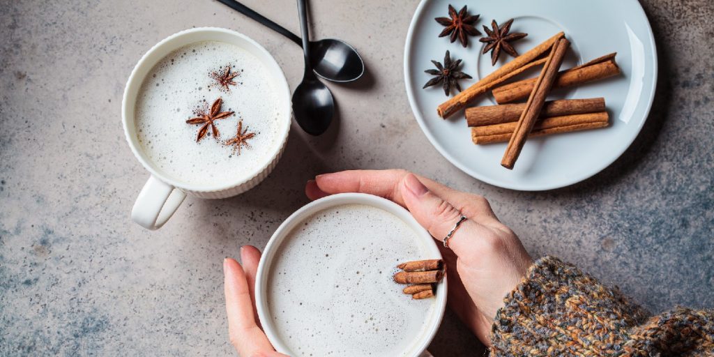 Homemade chai latte with cinnamon and star anise in white cup in hands, dark background, top view.